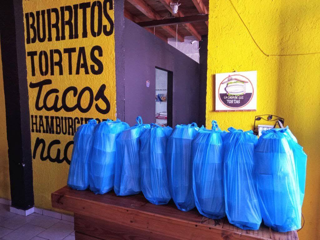Bolsas de un pedido grande alineadas en el mostrador, bajo el mural de burritos, tortas, tacos y hamburguesas